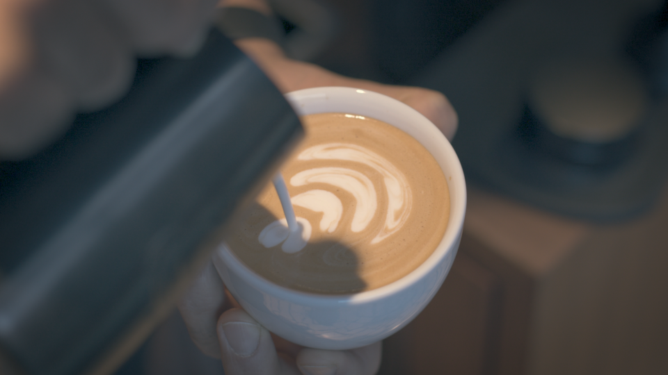 Aus einer Milchkanne wird Milch in eine kleine Tasse mit Kaffee gegossen und ergibt dort ein Muster