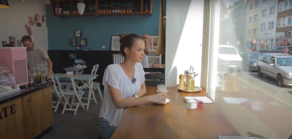 Eine junge Frau sitzt in einem Kaffee am Fenster, blickt nach draußen und hat vor sich einen Kaffee.