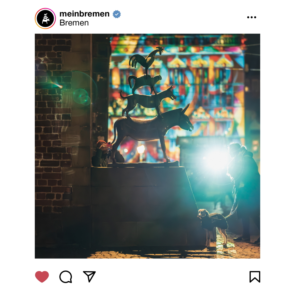 Das Foto zeigt die Bremer Stadtmusikanten von der Seite mit Blick zum Marktplatz. Im Hintergrund sind bunte Lichter zu sehen und vor der Statue steht eine Person mit einem Hund an der Leine.