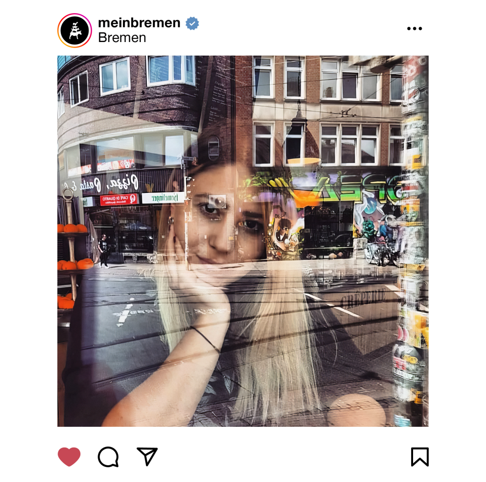 Das Foto zeigt eine Frau mit blonden Haaren, die hinter der Scheibe eines Cafés sitzt und nach draußen blickt. In der Schiebe spiegeln sich die Gebäude und die Straße der Bremer Sielwallkreuzung.