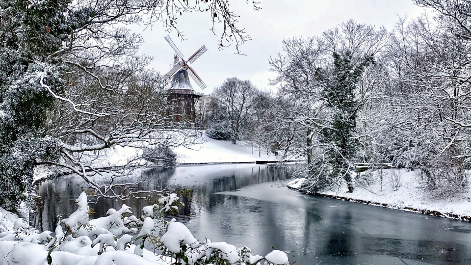 Eine eingeschneite Winterlandschaft ("Wallanlagen") mit einem zugefrorenem Wasserlauf. Im Hintergrund steht die eingeschneite "Mühle am Wall".