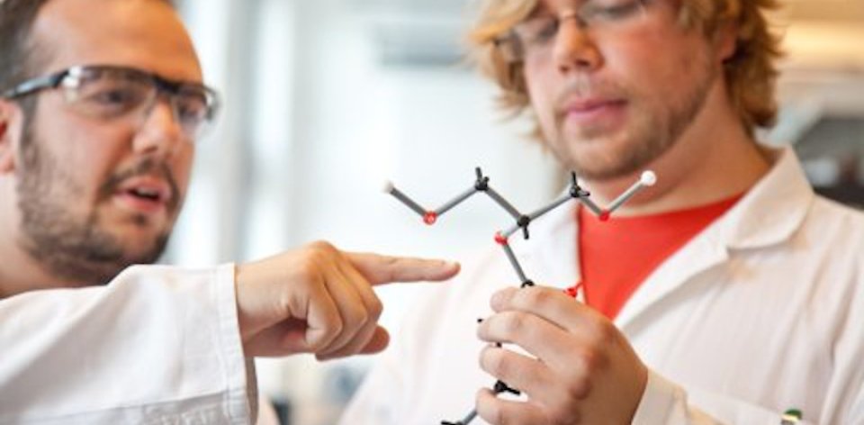 Zwei Männer in Schutzanzügen betrachten ein Modell einer chemischen Verbindung