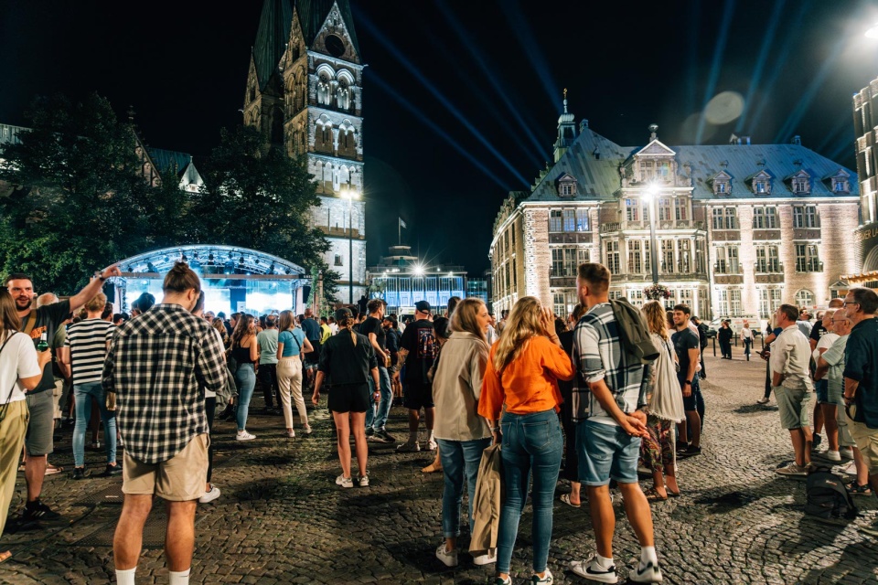 Groups of people stand in front of a large stage at night on the Domshof in Bremen.