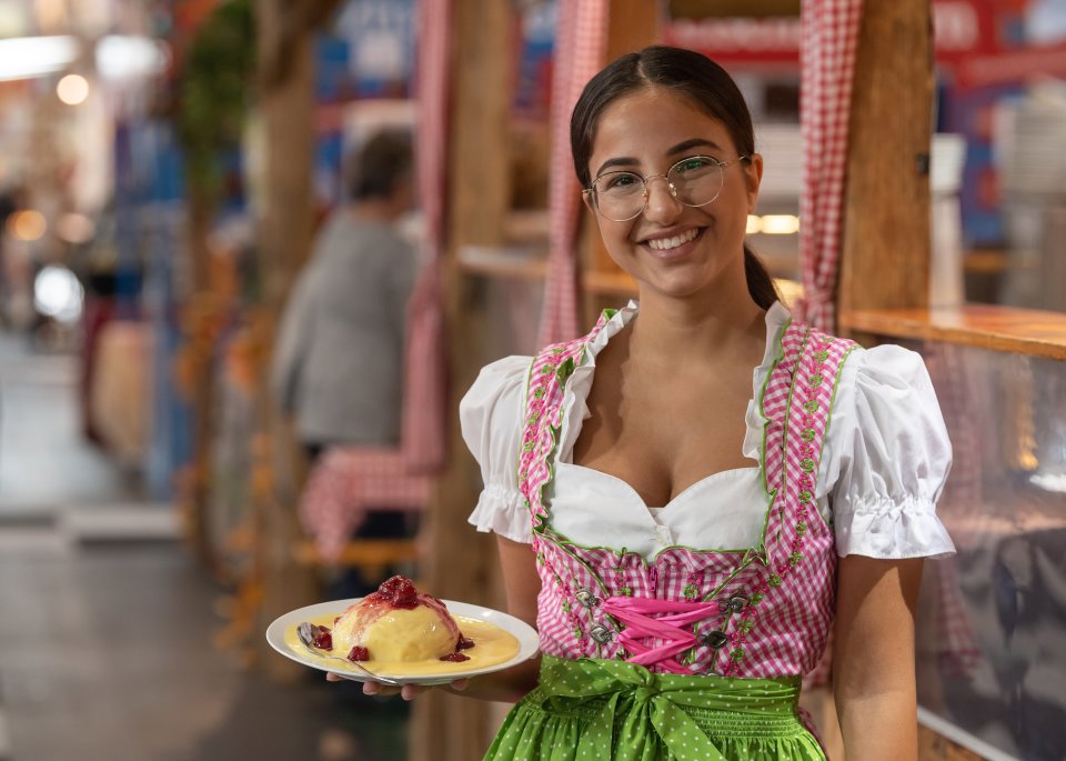 Eine junge Frau im Kleid hat einen Teller mit Essen in der Hand und grinst in die Kamera.