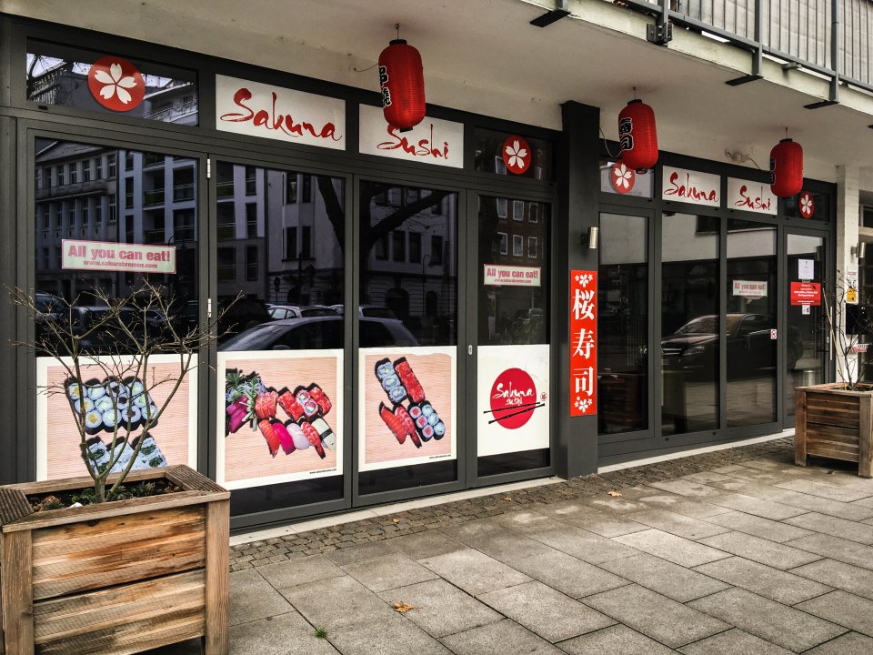Das Sushi-Restaurant Sakura von außen. Es stehen Blumenkübel dafür. Rote Laternen hängen von der Decke.
