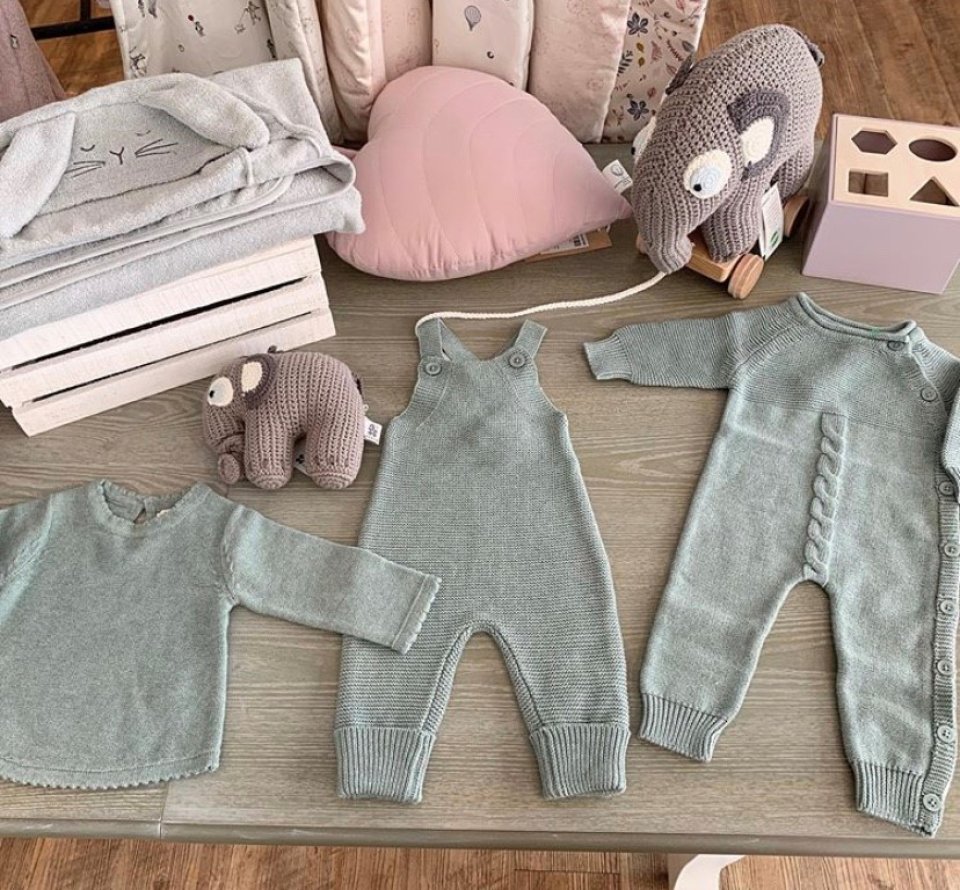 Die Aufnahme zeigt einen Pullover, eine Latzhose sowie einen Strampler für Babies. Zudem ist ein Kissen in Herzform sowie ein kleiner Stoff-Elefant zu sehen.