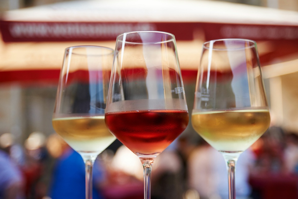 Zwei Weingläser mit einem roten und einem weißen Wein gefüllt stehen in einer Nahaufnahme vor dem Festgetümmel im Hintergrund auf einem Tisch.