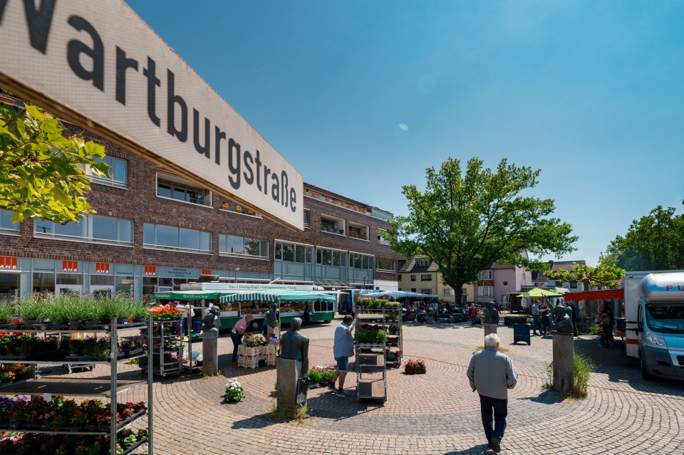 An einem Platz steht ein Schild auf dem Wartburgplatz steht. Darauf werden gerade Stände für einen Wochenmarkt aufgebaut.